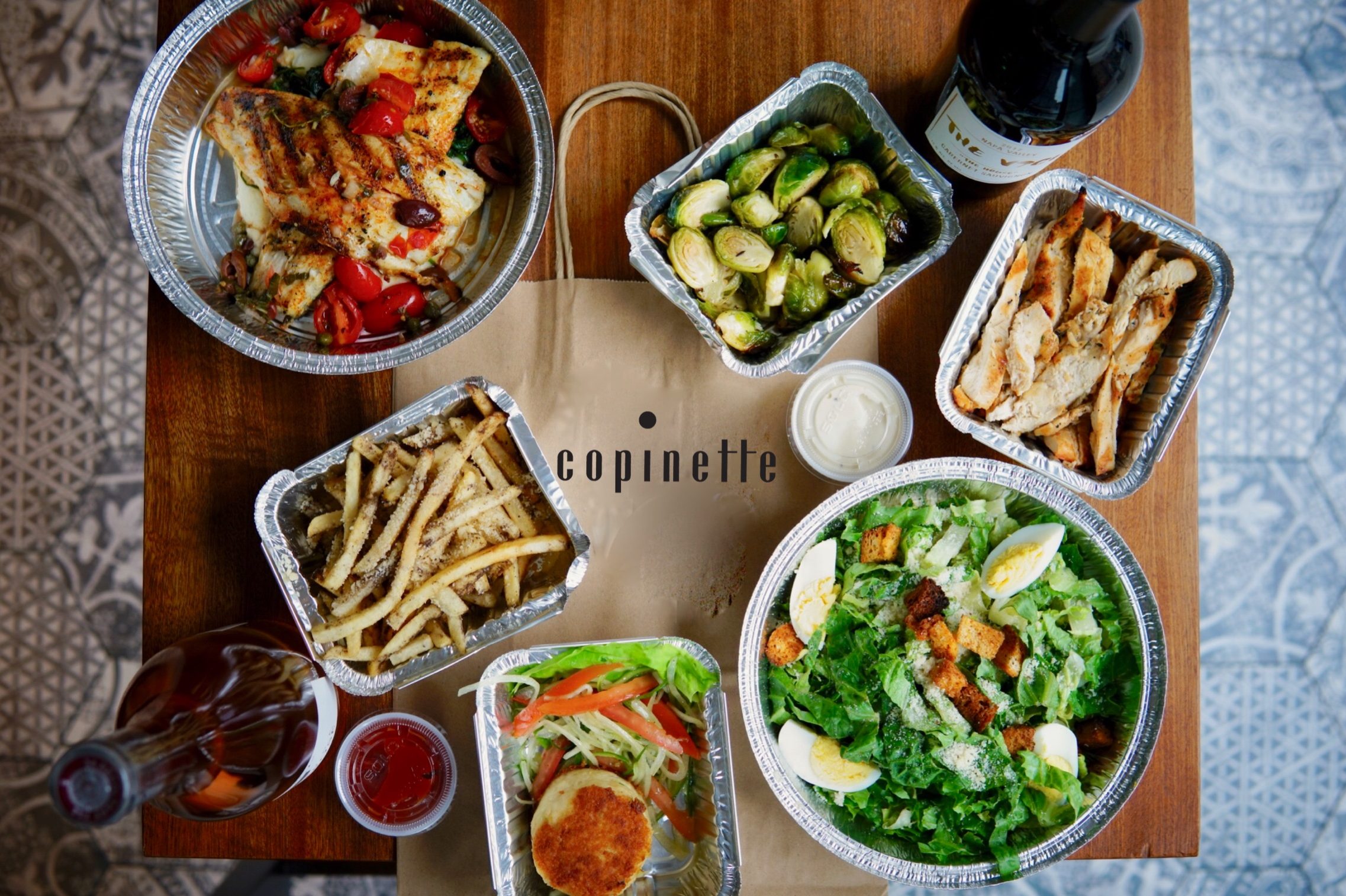 Overhead view of Copinette takeout with grilled chicken, salad, fries, and wine