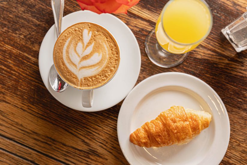 Latte with foam art, croissant on a plate, and glass of orange juice.