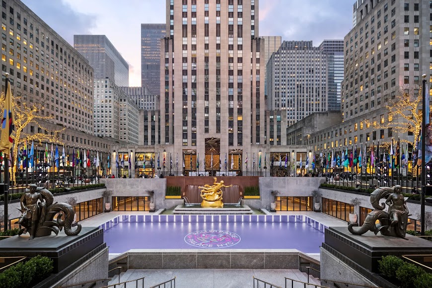 View of Rockefeller Center ice rink, flags, and Prometheus statue surrounded by skyscrapers and lights.