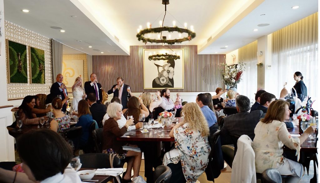 Guests toasting at tables in a bright dining room with chandelier and floral portrait on the wall.