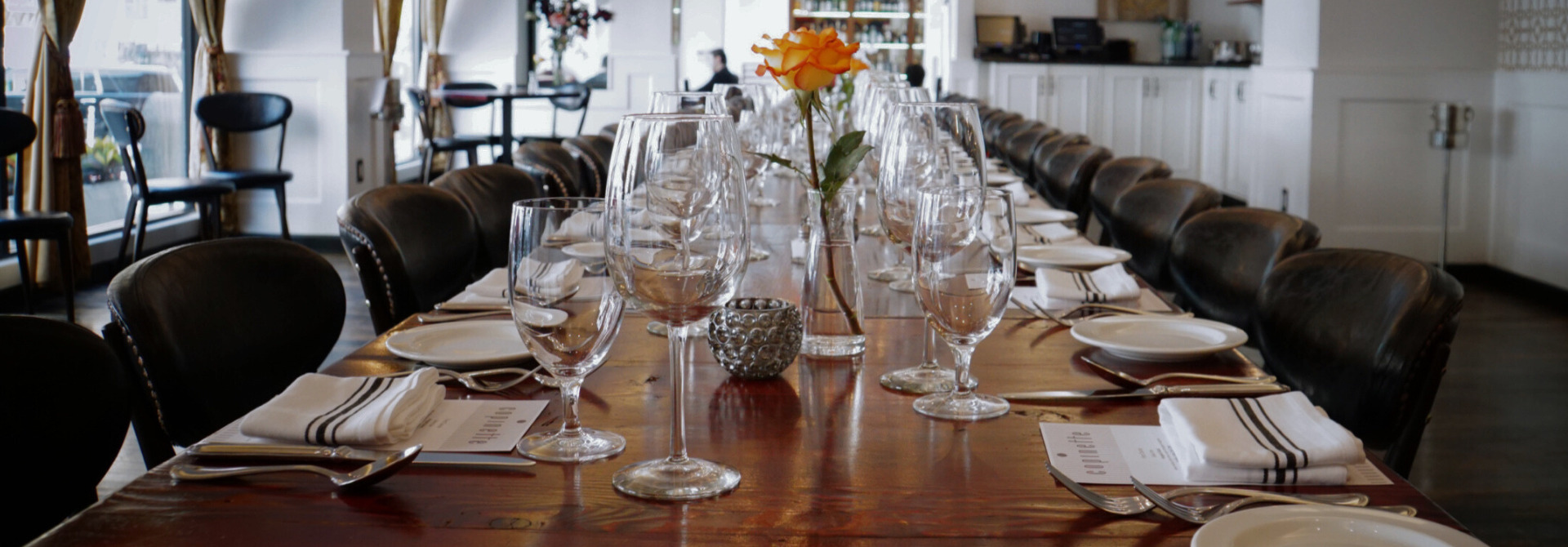Elegant long dining table set with glassware, plates, and a single orange rose centerpiece.