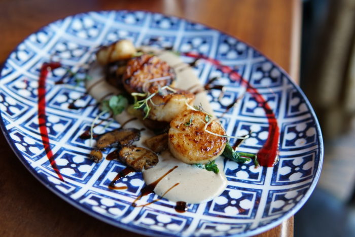 Seared scallops plated with mushroom purée, microgreens, and artistic sauce swirls on patterned dish.