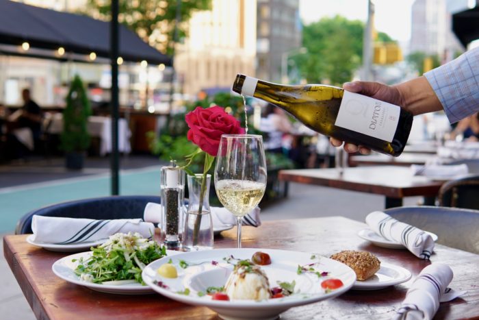 Server pouring white wine at an outdoor restaurant table set with salad and appetizers.