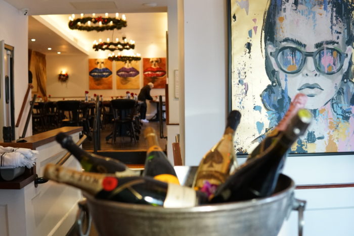 Champagne bottles in ice bucket near colorful portrait, with decorated dining room in background.