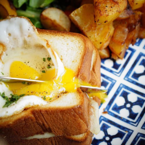 Runny egg on top of Croque Madame with breakfast potatoes and greens