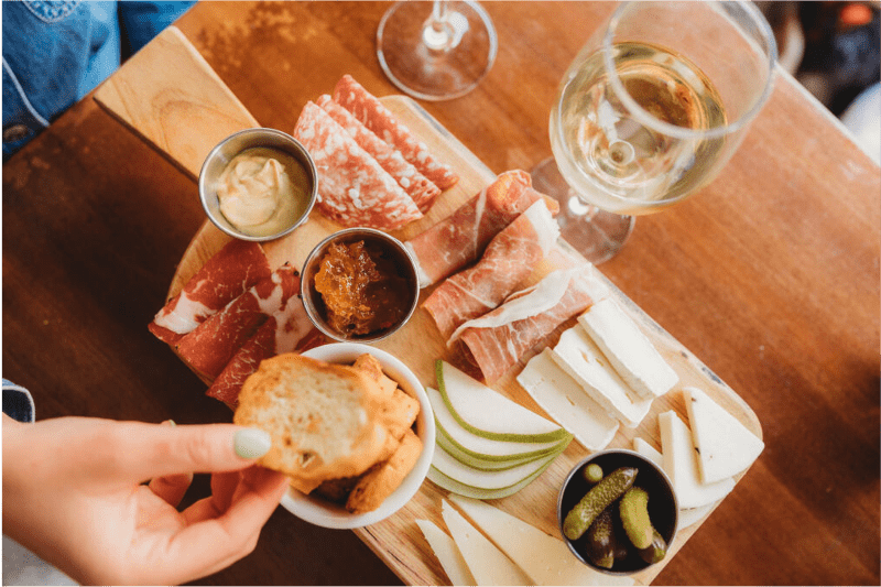 Hand reaching for bread on a charcuterie board with cheese, meats, accompanied with a glass wine