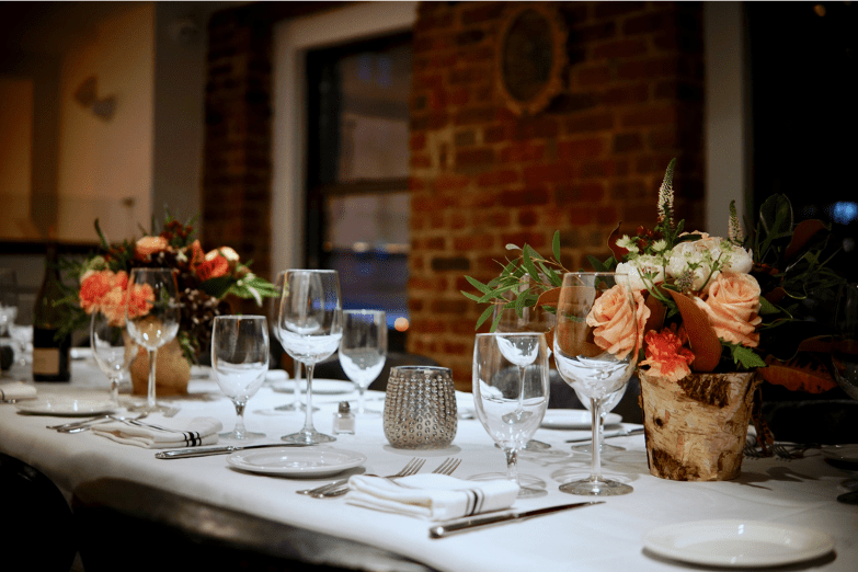 Close-up of a beautifully set private dining table with elegant glassware, floral centerpieces in rustic vases, and white tablecloth in a warm, brick-walled room