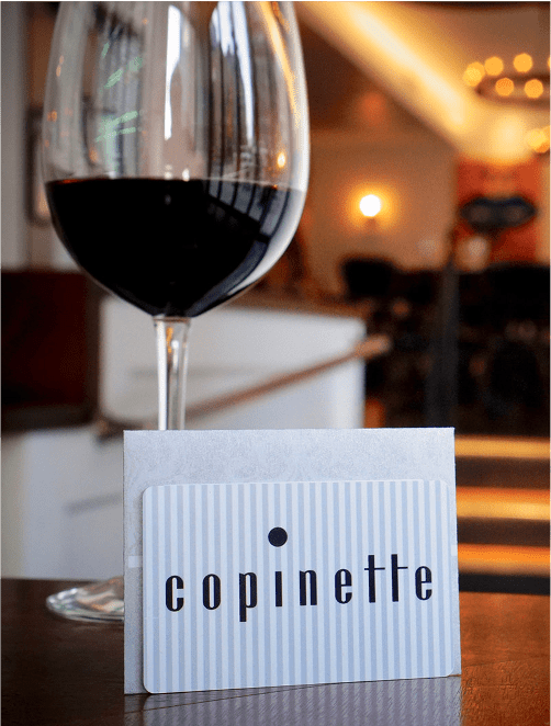 Glass of red wine next to a Copinette gift card in a white envelope, with a softly lit restaurant interior in the background