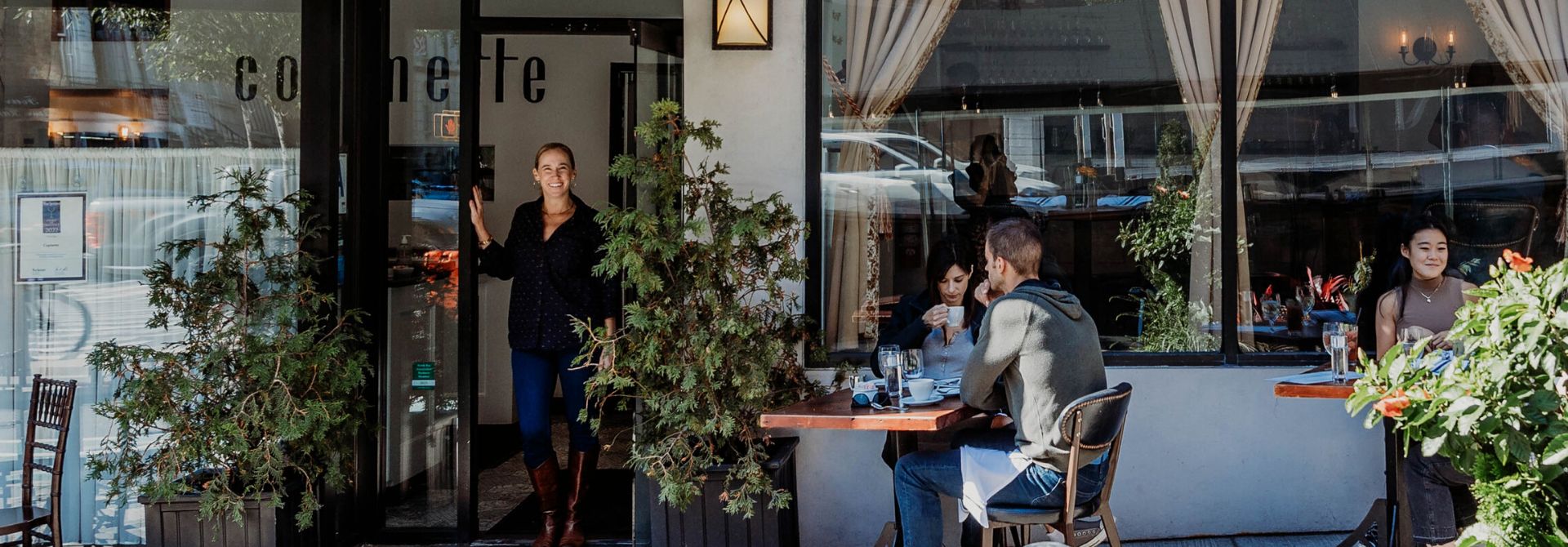 People sit at outdoor tables eating and drinking outside a cafe, while a woman stands in the doorway.