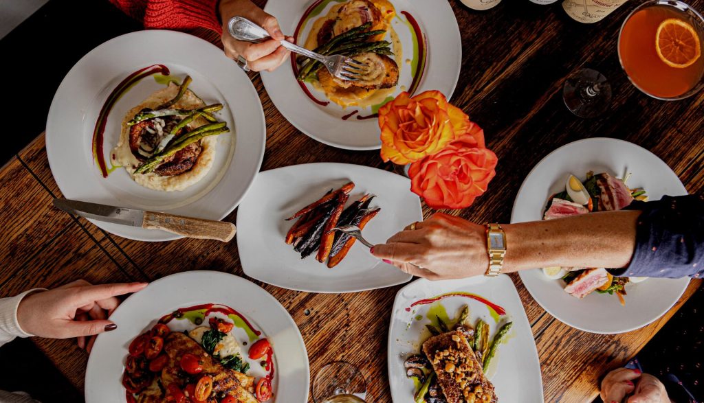 Top-down view of a rustic wooden table with multiple plated gourmet dishes, cocktails, and hands reaching to share food