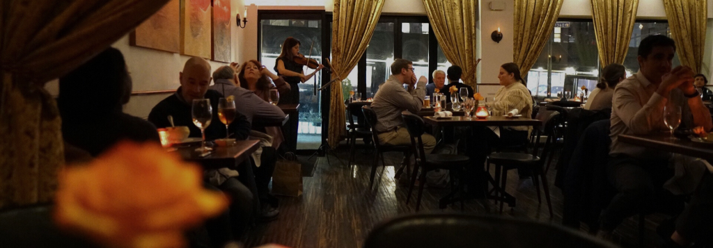 Candlelit restaurant dining room with guests enjoying dinner and live violin music near windows
