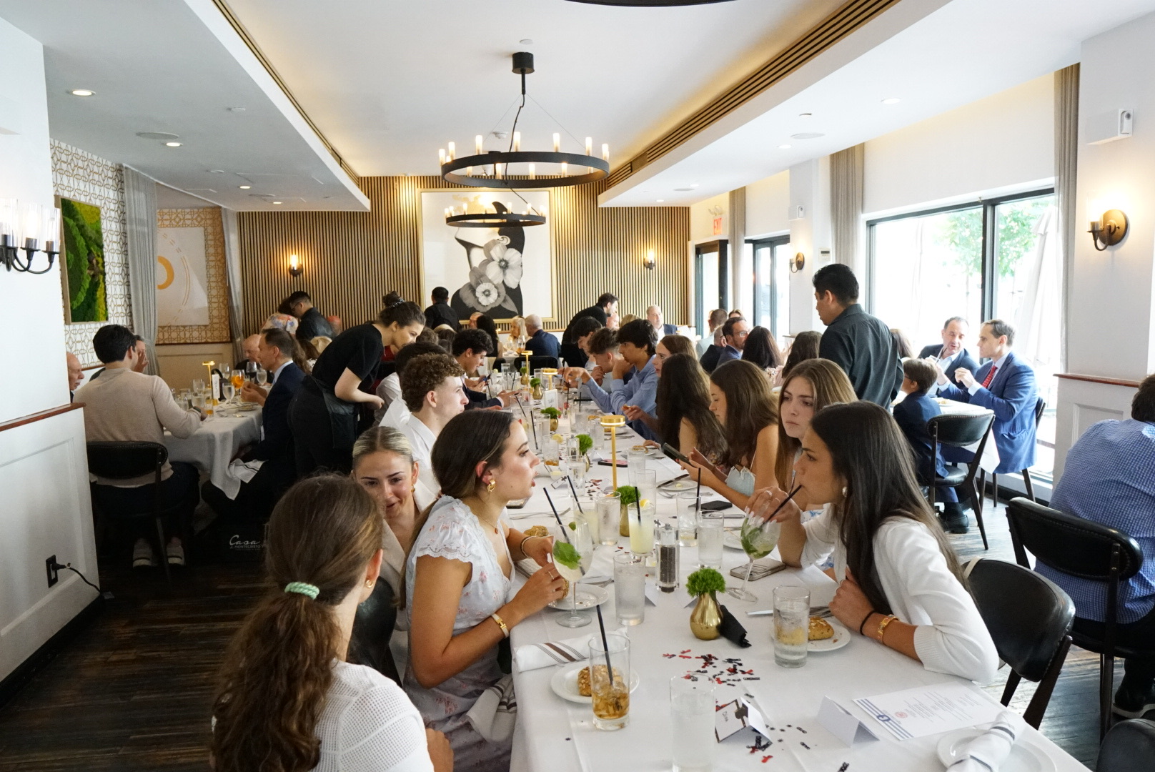 Large graduation party in a private dining room with guests seated at long tables, drinks and meals served, chandelier lighting, and tall windows.