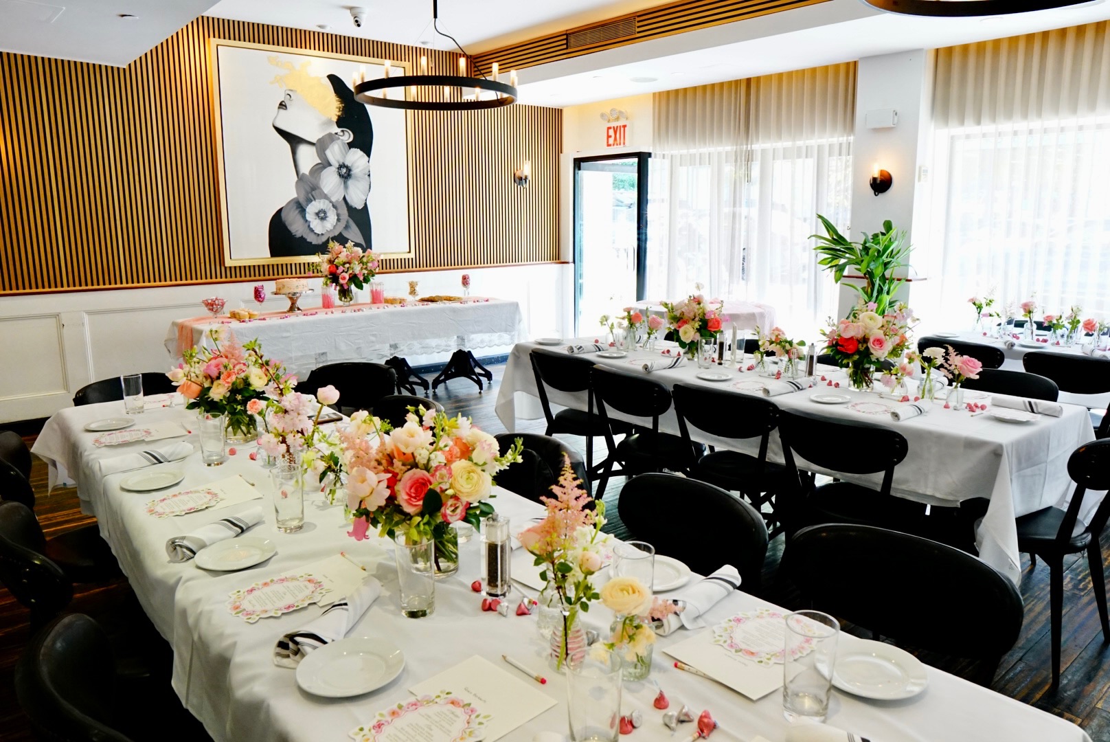 Private dining room decorated for a bridal shower with white-clothed tables, pastel floral centerpieces, black chairs, chandelier lighting, wood-paneled wall art, and tall windows.