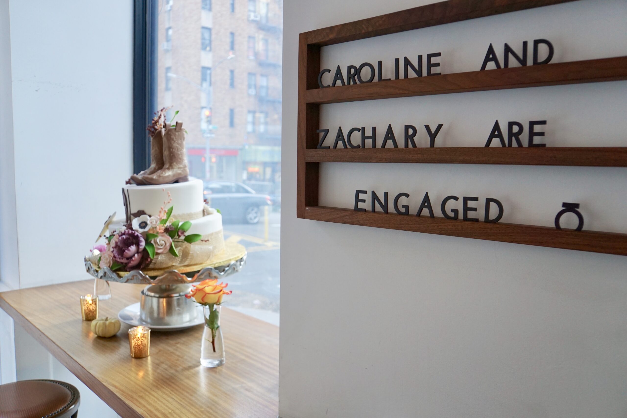 Engagement party display with tiered cake topped by cowboy boots, floral accents, candles, and wall sign reading Caroline and Zachary are engaged.
